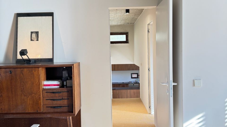 Minimalist interior room with a raw concrete ceiling, light gray walls, and carpeted floor, featuring a mid-century wooden cabinet and a white chair. An open white door leads to a hallway with built-in wood cabinetry and a small horizontal window, with sunlight casting striped shadows on the wall.