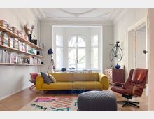 Bright living room with ornate ceiling moldings and a large bay window, featuring a mustard-yellow sofa, patterned rug, and a leather lounge chair. Wall-mounted bookshelves line one side, and a bicycle hangs above a wooden chest near a doorway.