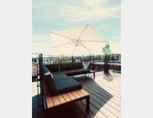 Moderne Dachterrasse mit Holzboden, schwarzer Sitzgruppe aus Polstern und einem hellen Sonnenschirm. Im Hintergrund sind Geländer mit Verzierungen und Gebäude in einer städtischen Umgebung sichtbar.
