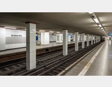 U-Bahn-Station mit der Beschriftung „Potsdamer Platz“, bestehend aus einem langen, rechteckigen Bahnsteig, flankiert von Gleisen. Weiße Fliesenwände und rechteckige Säulen prägen das schlichte, beleuchtete Innendesign, während ein Zug am rechten Gleis sichtbar ist.