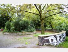 Ein kleiner Parkbereich mit einer runden, niedrigen Steinmauer, die ein bepflanztes Beet umschließt. Im Vordergrund steht ein großer Baum, umgeben von dichtem Grün und weiteren Bäumen im Hintergrund.