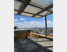 Rooftop terrace with large gray floor tiles, metal railings, and a slatted pergola roof casting striped shadows. Beyond the edge is a wide city skyline under a blue sky with scattered clouds, including a tall TV tower in the distance.