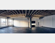 Weitläufiger, leerer Raum mit Sichtbetondecke und großen Fenstern auf der linken Seite, die einen Blick auf Außengebäude ermöglichen. Die weiße Wand rechts enthält eine geschlossene Garagentür, eine kleine Küchenzeile sowie eine Feuerschutzausrüstung.