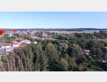 Luftaufnahme eines ländlichen Gebiets mit dicht bewaldeten Flächen im Vordergrund, kleinen Häusern mit roten und weißen Dächern in der Mitte und offenen Feldern sowie einem See in der Ferne. Der Himmel ist klar und blau.