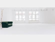 Ein heller, minimalistisch gestalteter Raum mit weißen Wänden und weißem Boden, großen Fenstern auf der gegenüberliegenden Seite, die viel Licht hereinlassen, einer dunkelgrünen Chesterfield-Couch auf der linken Seite und einem dünnen weißen Baumstamm als Dekoration.
