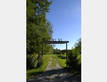 Eingang zu einem Naturgebiet mit einem Holzschild, auf dem "Kahnfährhafen Neu Zauche" steht, umgeben von grünen Bäumen und Wiesen. Im Hintergrund sind kleine Holzkonstruktionen und ein schmaler Weg sichtbar.