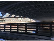 Blick unter eine moderne Metallbrücke mit gebogenen Trägern hindurch, im Hintergrund ist ein mehrstöckiges Bürogebäude mit einer Fassade aus Glas und hellen Paneelen zu sehen. Darunter fließt ein Wasserkanal, der teilweise im Schatten der Brücke liegt.