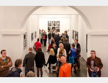 Innenansicht einer Galerie mit weißen Wänden, die mit Schwarz-Weiß-Fotografien dekoriert sind. Viele Menschen füllen den Raum, betrachten die Bilder oder stehen in Gruppen zusammen und unterhalten sich, während ein großer Bogen im Vordergrund den Raum umrahmt.