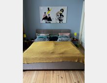 Schlafzimmer mit einem grauen Polsterbett, bedeckt mit einer senfgelben Decke und mehreren farbigen Kissen. Über dem Bett hängt ein großes Bild mit abstrakten Figuren, und auf beiden Seiten des Bettes stehen Nachttische mit gelben Tischlampen.