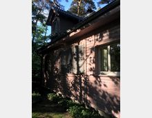 Holzhaus mit rosa Fassade, Klappläden an den Fenstern und einem schrägen Dach, umgeben von hohen Bäumen, die Schatten auf die Wände werfen. Im Vordergrund sind Pflanzen und Sträucher zu sehen.