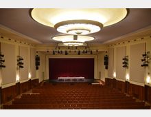 Ein Veranstaltungsraum mit einer Bühne, roten Vorhängen und einer Reihe von vier Tischen mit Stühlen. Der Raum hat beige Wände, mehrere Wandlampen, hölzerne Wandverkleidungen und eine große Deckenbeleuchtung, mit Reihen von braunen Sitzplätzen davor.