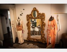 Raum mit einem großen goldenen Spiegel in kunstvollem Rahmen, flankiert von zwei Schaufensterpuppen in traditionellen Kimonos. Im Spiegel spiegelt sich ein Tisch mit weißen Stühlen und ein grüner gefliester Ofen; daneben sind eine offene Tür, eine Lampe und ein Teppich mit Leopardenmuster zu sehen.