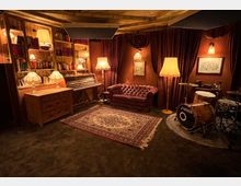 Gemütlicher Raum mit dunklen Vorhängen, Regalwänden voller Bücher und mehreren Steh- und Tischlampen. Im Vordergrund ein dekorativer Teppich, ein Chesterfield-Sofa, ein Schlagzeug und ein Klavier; die Einrichtung wirkt im klassisch-vintage Stil gehalten.