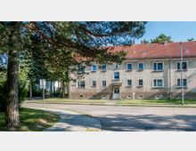 Wohngebäude mit zwei Stockwerken in einer Wohngegend, erkennbar an der hellbraunen Fassade, einem rot gedeckten Satteldach und mehreren Fenstern. Im Vordergrund befinden sich eine Straße, ein Gehweg sowie Bäume, die Schatten werfen.