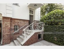 Backsteinhaus mit hellem Putz an der oberen Fassade, einer halbhohen Treppe mit verziertem, weißem Geländer und einer Tür im oberen Bereich. Links ein kleines Fenster mit Metallsprossen, rechts ein gepflegter Hecken- und Buschbereich.