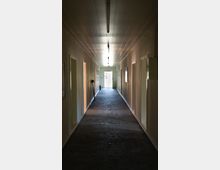 Ein schmaler, langer Flur mit weißen Wänden und einer dunklen Teppichbodenoberfläche, beleuchtet durch deckenmontierte Leuchtstofflampen. Der Flur führt zu einer hell beleuchteten Tür am Ende, mit mehreren seitlichen Türen auf beiden Seiten.