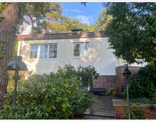 Ein weißes Einfamilienhaus mit einem flachen Dach, teilweise verkleidet mit rotem Klinker, umgeben von dichtem Grün und hohen Bäumen. Im Vordergrund befindet sich ein gepflasterter Weg mit zwei Laternen, die von Sträuchern und Pflanzen eingerahmt werden.