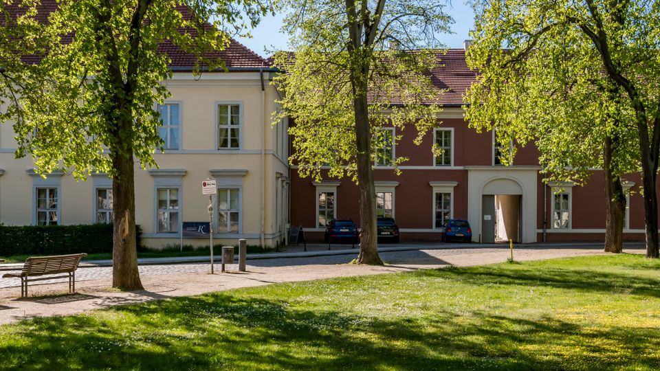 Tree-lined courtyard with a grassy lawn and benches in the foreground, facing a row of low historic buildings with red tile roofs and evenly spaced windows. A cobblestone path and small parking area run along the buildings, including a central arched entryway.