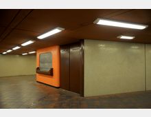 U-Bahn-Passage mit orangefarbenen und braunen Wänden, einer geschlossenen Rolltür in einem orangefarbenen Kioskbereich und gefliesten Böden. Weiße, rechteckige Leuchtstofflampen sind gleichmäßig in der braunen Decke angebracht.