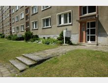 Mehrstöckiges Wohngebäude mit einer hellbraunen Fassade, rechteckigen Fenstern und einem Hauseingang mit Nummer „86“. Vor dem Gebäude befinden sich ein kleiner Gehweg, eine Grünfläche mit niedrigen Pflanzen und Sträuchern sowie ein Briefkastenstand.