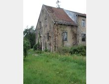 Altes, zweistöckiges Gebäude mit bröckelnder Fassade und Ziegeldach, teilweise von Ranken und Kletterpflanzen bedeckt. Das Gebäude steht inmitten eines überwucherten Gartens mit grasbewachsenem Boden und vereinzelten Büschen.