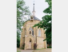 Kirchengebäude aus gelbem Backstein mit gotischen Fenstern und spitzem Turm, umgeben von Bäumen und blühenden Rosensträuchern.