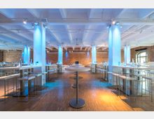 Großer Veranstaltungsraum mit modernen, hohen Stehtischen und Hockern auf einem Holzboden. Die Backsteinwände und weißen Deckenträger sind blau beleuchtet, und große Rundbogenfenster lassen Tageslicht herein.