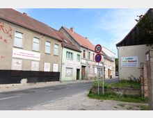Straßenszene in einem Wohngebiet mit älteren Häusern, teilweise mit renovierungsbedürftigen Fassaden. An den Gebäuden und auf einem Plakat an der Wand sind Protestbotschaften zu sehen, die gegen Schwerlastverkehr und für eine Verkehrsberuhigung plädieren; Verkehrsschilder zeigen eine Geschwindigkeitsbegrenzung von 30 km/h und ein Halteverbot an.