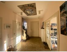 Ein Eingangsbereich in einem Haus mit hellen Wänden, einer verzierten Deckenmalerei, und einem Steinboden. Links führen Treppen nach oben, rechts befindet sich eine Glastür, die in ein Arbeitszimmer mit Schreibtisch und Regalen führt.