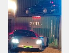 Ein silbernes Sportwagenmodell mit eingeschalteten Scheinwerfern steht vor einem Stapel Schiffscontainern. Auf dem oberen Container ist ein schwarzes Auto abgestellt, der Container trägt den Schriftzug "CAPITAL".