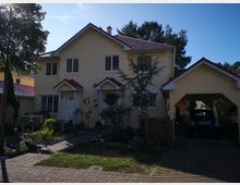Zweistöckiges gelb verputztes Wohnhaus mit rotem Satteldach, flankiert von einem angelegten Gartenbereich und einer Einfahrt mit Carport, unter dem ein Auto parkt. Im Hintergrund sind Bäume und blauer Himmel zu sehen.