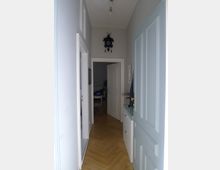 Ein schmaler Flur mit hellgrauen Wänden und Parkettboden. Im Hintergrund ist eine geöffnete Tür zu einem weiteren Raum sichtbar, an den Wänden hängen eine Uhr und Bilder, und rechts steht eine Kommode.