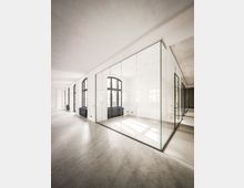 Heller, minimalistischer Innenraum eines modernen Büro- oder Ateliergebäudes mit poliertem Betonboden, weißen Wänden und hohen, bogenförmigen Fenstern entlang der linken Seite. In der Mitte steht ein gläserner Kubus als abgetrennter Raum, dessen transparente Wände Licht und Fensterreflexionen zeigen.