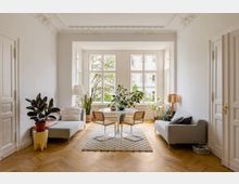 Helles Wohnzimmer mit hohen Decken, verzierten Stuckdetails und einem großen Fensterbereich, der viel Tageslicht hereinlässt. Der Raum ist mit zwei Sofas, einem runden Tisch mit Stühlen und zahlreichen Zimmerpflanzen dekoriert, der Boden besteht aus Holzparkett.