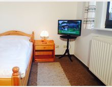 Ein schlicht eingerichtetes Schlafzimmer mit einem Einzelbett aus hellem Holz, einem passenden Nachttisch mit Tischlampe, einem kleinen Fernseher auf einem schwarzen Ständer und einem Fenster mit dekorativ gemusterten Vorhängen. Der Raum ist mit hellem Teppichboden und einer Heizung an der Wand ausgestattet.