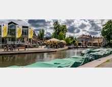 Ein Flussufer mit überdachten Booten im Vordergrund, flankiert von einer Promenade mit Restaurants, Cafés und gelben Sonnenschirmen. Im Hintergrund sind moderne und traditionelle Gebäude sowie grüne Bäume zu sehen.