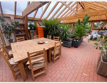 Ein lichtdurchfluteter Raum mit einem Holzdach und großen Glasflächen, eingerichtet mit einem rechteckigen Holztisch und mehreren Holzstühlen. Umgeben von zahlreichen Topfpflanzen, befindet sich im Hintergrund ein auffälliger orangefarbener Retro-Kühlschrank und Regale mit Dekorationen.