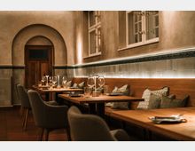 Gemütlicher Restaurant-Innenraum mit Holz­tischen, gepolsterter Sitzbank mit Kissen und Stühlen, jeweils mit Gläsern und Besteck gedeckt. Helle Putzwände mit Fliesenbordüre, hohe Fenster und ein Rundbogen mit Holztür prägen den Raum.