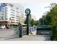 Eingang zur S-Bahn-Station "Anhalter Bahnhof" in einer städtischen Umgebung, mit einem markanten "S"-Symbol auf einem Pfeiler. Im Hintergrund sind moderne Gebäude, eine Straße mit Autos und Fahrräder im Vordergrund zu sehen.