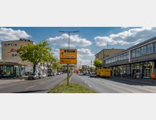 Straßenszene mit mehrspuriger Fahrbahn, flankiert von Gebäuden mit Geschäftsfassaden, darunter ein Optikergeschäft und ein DHL-Fahrzeug. Ein gelbes Verkehrsschild in der Mitte zeigt Richtungen zum Zentralen Festplatz und nach Wedding an, während der Himmel teils bewölkt ist.