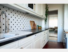 Helle Küche mit weißen Schränken, schwarzer Arbeitsplatte und einer Spüle mit verchromtem Wasserhahn. Der Fliesenspiegel hat ein schwarz-weißes Muster, und im Hintergrund führt eine Türöffnung in ein Wohnzimmer mit Sofa.