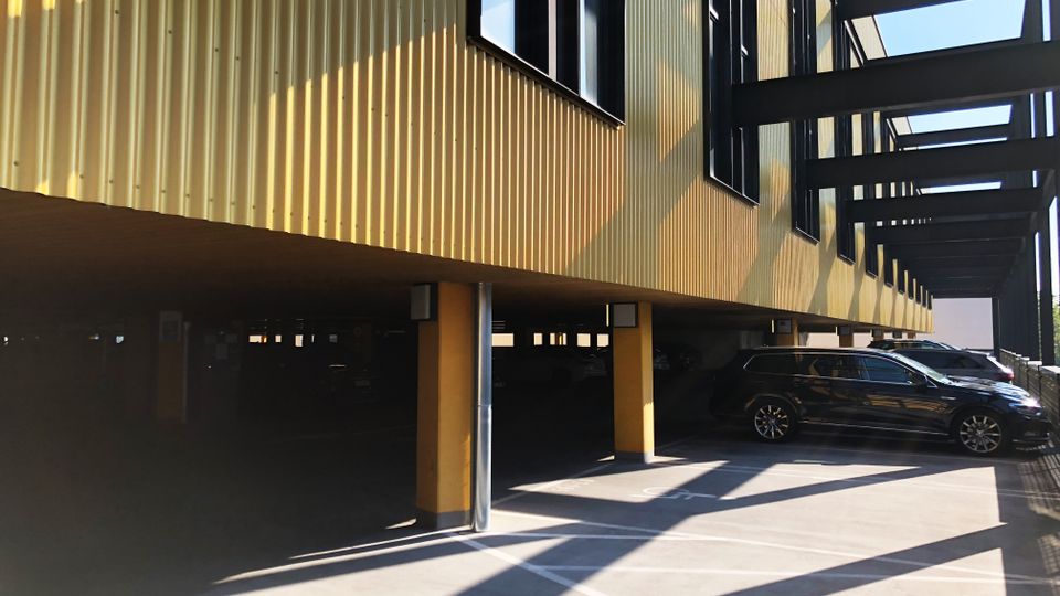 Eine Parkgarage mit gelber, wellenförmiger Fassade und freistehenden Pfeilern, unter einem modernen Gebäude mit Glasfenstern und schwarzem Stahlgerüst. Im Vordergrund sind schattige Parkplätze sichtbar.