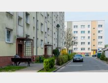 Mehrstöckige Wohnhäuser in einer städtischen Wohnsiedlung, mit gelblichem und weißem Putz sowie roten Klinkerbereichen an den Eingängen. Im Vordergrund ist eine parkähnliche Anlage mit einem Gehweg, einer Bank, Laternen und Sträuchern zu sehen, rechts stehen geparkte Autos.