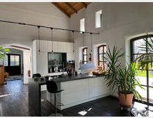 Eine moderne, helle Küche mit weißen Einbauschränken und einer dunklen Kochinsel, darüber hängen minimalistische Pendelleuchten. Große Fenster mit schwarzen Rahmen lassen viel Tageslicht herein und bieten einen Blick auf einen Garten, während Pflanzen und Holzelemente den Raum gemütlich gestalten.