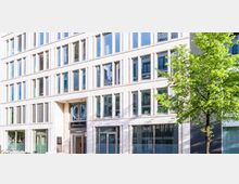 Moderne Bürogebäude-Fassade mit heller Steinverkleidung und großen Fenstern in regelmäßiger Anordnung; im Vordergrund ein grüner Baum und einige geparkte Fahrräder.