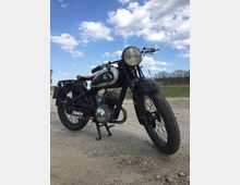 Ein klassisches schwarzes Motorrad mit gerade aufgebocktem Ständer steht auf einer unbefestigten Landstraße. Im Hintergrund sind Grasflächen, Bäume und ein blauer Himmel mit weißen Wolken zu sehen.
