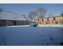 Verschneiter Bauernhofhofplatz mit einem Fachwerkhaus mit schneebedecktem Satteldach links und einem langen Backstein-Stallgebäude mit Holztoren rechts. Im Vordergrund liegt eine weite Schneefläche mit wenigen Grashalmen, dahinter stehen kahle Bäume vor blauem Himmel.