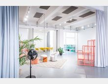 Heller, offener Innenraum eines modernen Studios oder Coworking-Spaces mit hohen Decken, Akustikpaneelen und Vorhängen als Raumteiler. In der Mitte stehen Sitzmöbel auf einem Teppich, dahinter ein gelber Tischbereich, Pflanzen, ein Regal und rote Gitterpodeste neben großen Fenstern.