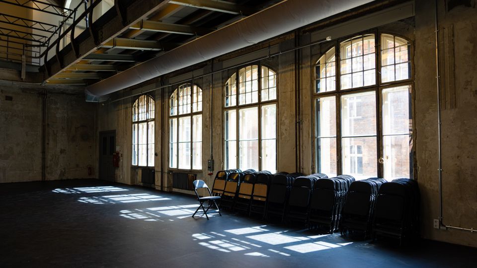 Großer, leerer Industriehallenraum mit rauen Betonwänden und hoher Decke mit sichtbaren Stahlträgern und Lüftungskanal. Entlang einer Wand stehen hohe, bogenförmige Sprossenfenster, durch die Licht auf den Boden fällt; darunter sind gestapelte Klappstühle angeordnet.
