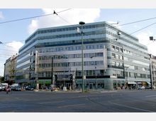 Ein moderner Büro- und Einkaufsgebäudekomplex am U-Bahnhof Schönhauser Allee in Berlin, mit einer Fassade aus Glas und Metall; im Vordergrund eine Straßenkreuzung mit Straßenbahnlinien und Verkehrsschildern.